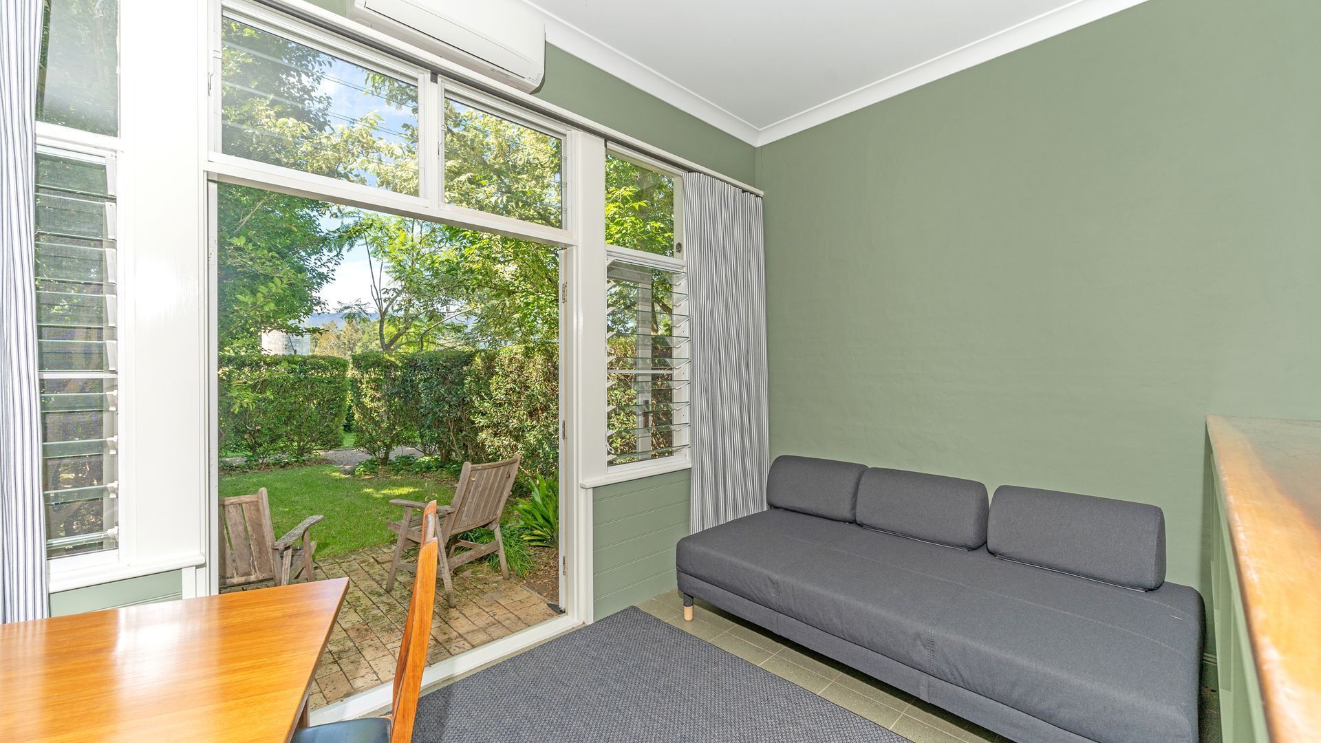 Ein Zimmer mit grauem Sofa, einem Holztisch und Blick auf einen Garten durch große Fenster.