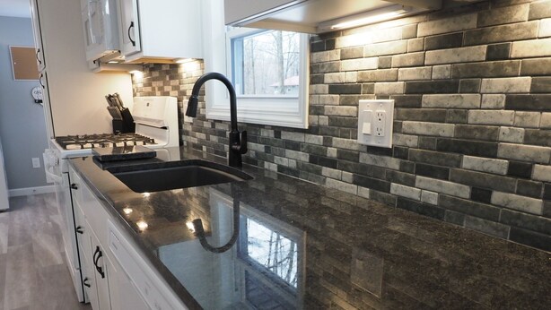 A modern kitchen with a granite countertop, a stainless steel sink, and a brick backsplash. - The Red Haven at Conneaut Lake (Conneaut Lake)