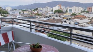 A balcony with a table and chairs, a potted plant, and a view of a cityscape with buildings and mountains in the distance. - Depto Con Inmejorable Ubicación (Salta)