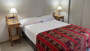 A neatly made bed with white linens and a red patterned bedspread, flanked by two wooden nightstands with lamps, in a room with plain walls and a tiled floor. - Depto Con Inmejorable Ubicación (Salta)