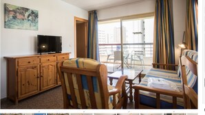 A hotel room with a wooden cabinet, a television, a striped sofa, a dining table, and a balcony with curtains. - AQUARIUM II 3-3 (benidorm)