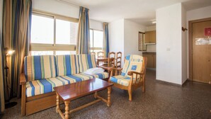 Una sala de estar con un sofá a rayas, una mesa de centro de madera y un sillón de madera. Se ve un área de cocina al fondo.