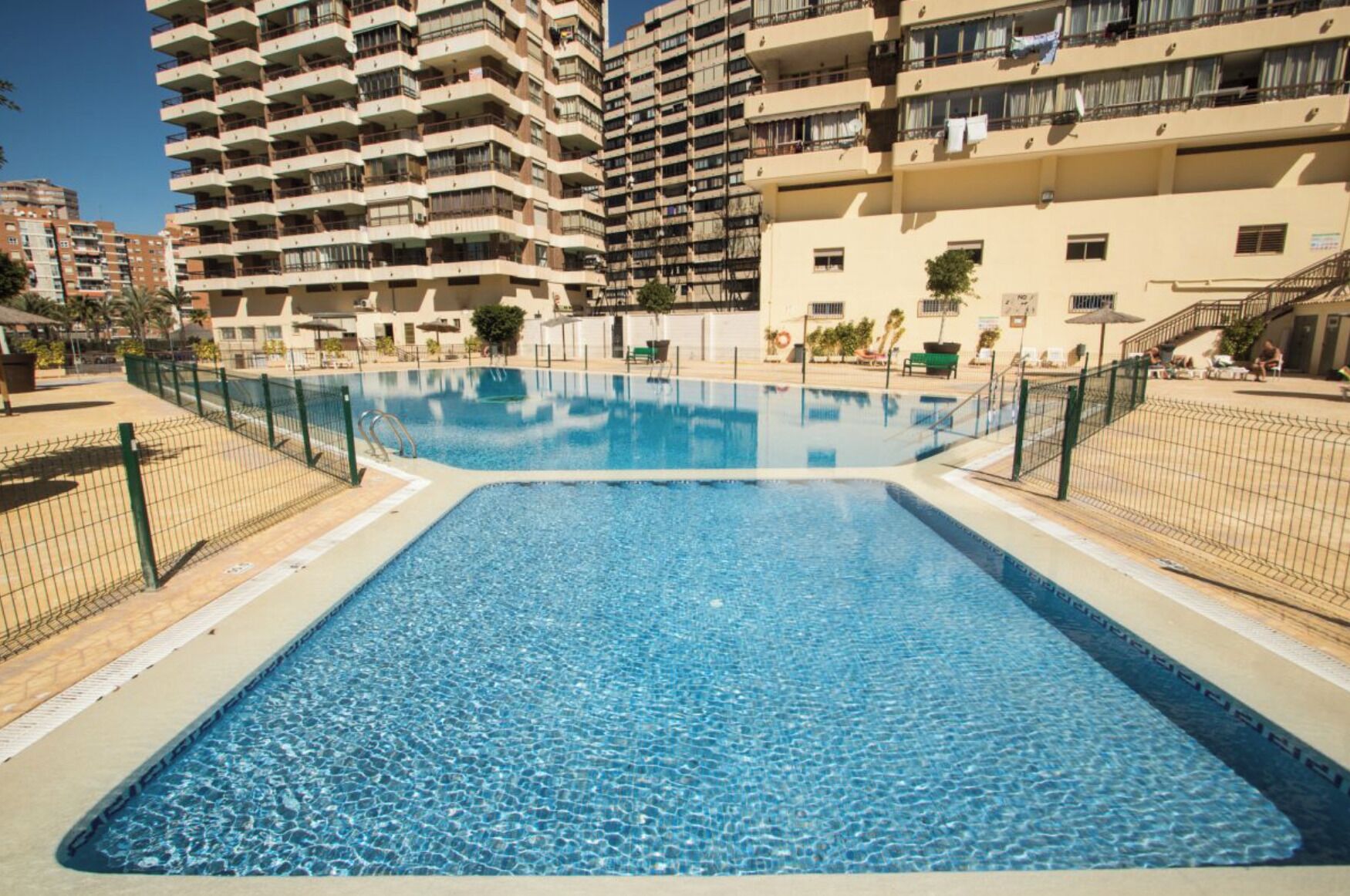 A rectangular swimming pool with a surrounding fence, set in a residential area with high-rise apartment buildings in the background.