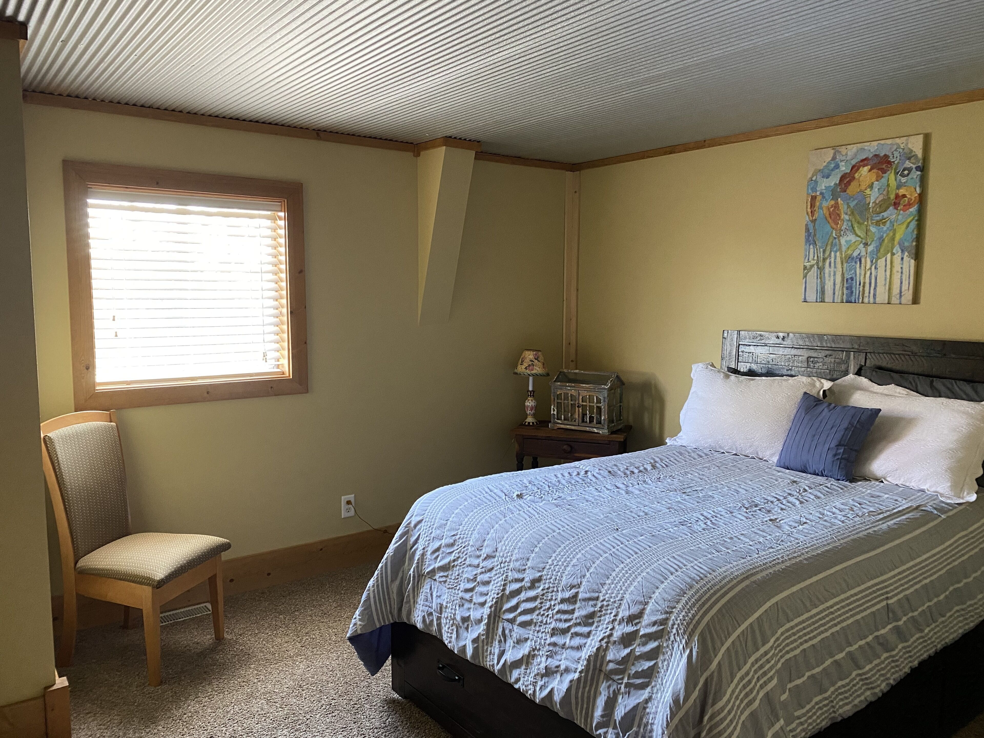 Une chambre à coucher avec un lit, une chaise, une fenêtre avec des stores, une table de chevet avec une lampe et un tableau au mur.