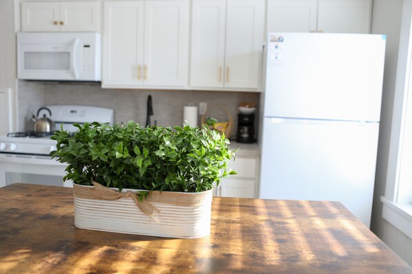 Una cocina con armarios blancos, microondas, hervidor, refrigerador y una mesa de madera con planta en maceta.