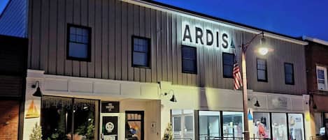 Un edificio con un cartel que dice 'ARDIS' en la fachada, escaparates con señalización visible y un asta con la bandera estadounidense.