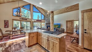 A spacious kitchen with a large island, wooden cabinets, and a stone fireplace. There are large windows offering views of the outdoors.