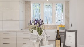 Una cocina con armarios blancos, un jarrón de flores de lavanda, una botella de vino, un letrero de bienvenida enmarcado y una tabla de cortar con galletas.