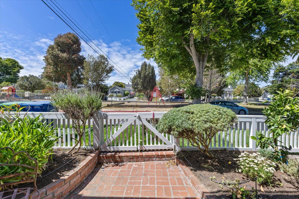 A residential area with a brick pathway leading to a white picket fence, surrounded by greenery and trees.