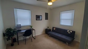 A small, well-lit room with a desk, chair, and sofa. There are two windows with blinds, a ceiling fan, and a small plant on the desk.