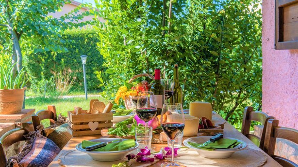 Spisebord udendørs dækket med tallerkener, glas og en flaske, omgivet af grøn vegetation og en træstol.
