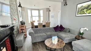 A modern living room with a fireplace, grey sofas, and a dining area with natural light. - Sea views just yards from restaurants, pubs & shops. Sleeps 16-18, 8 x en-suite. (Tintagel)