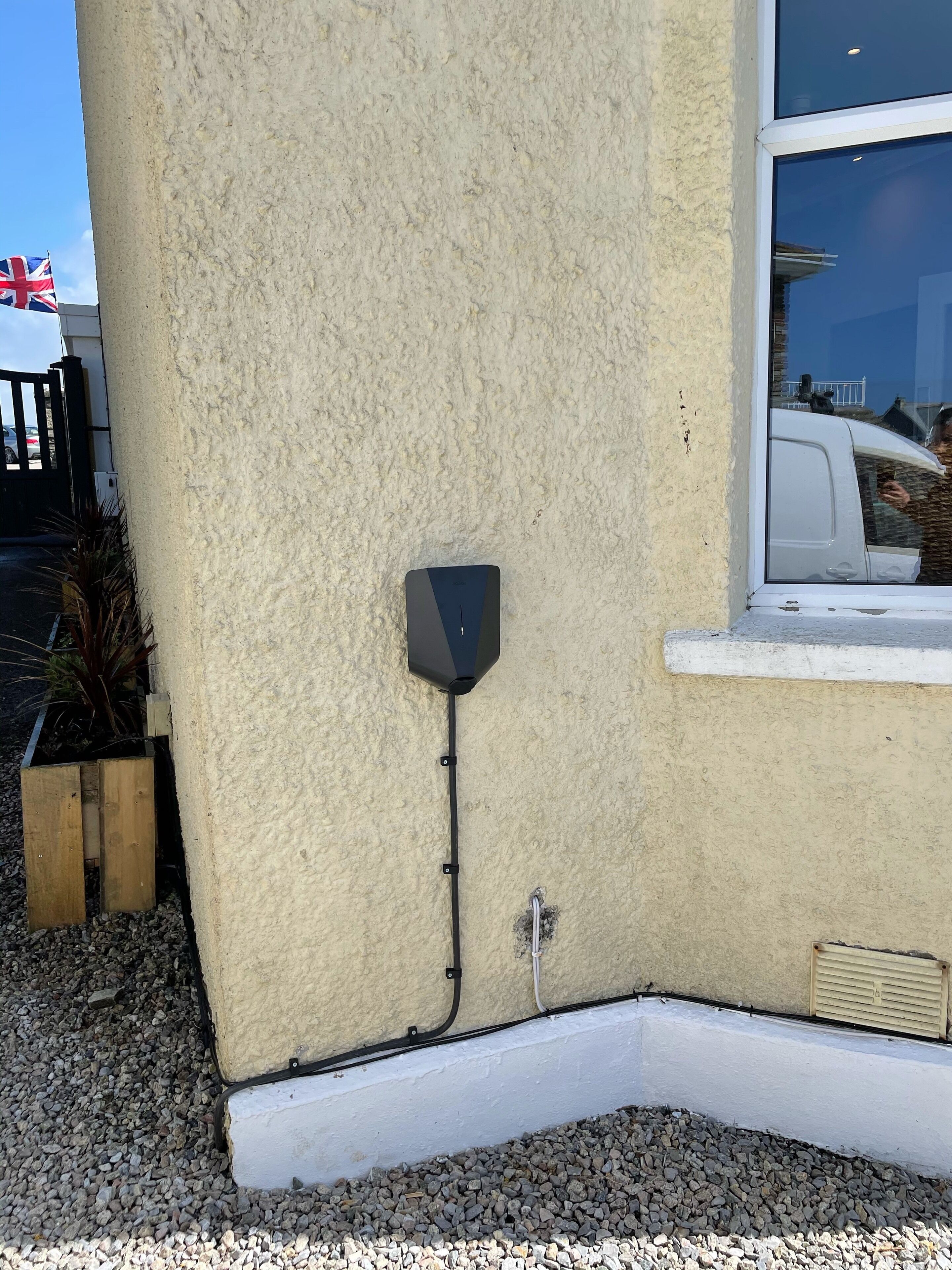 Una esquina de edificio con un muro beige texturizado, una ventana con marco blanco y una cámara de seguridad negra fijada en la pared.