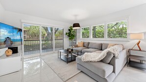 Una sala de estar moderna con un sofá gris, una mesa de centro y un televisor. Amplias ventanas con persianas ofrecen vistas a la vegetación exterior.