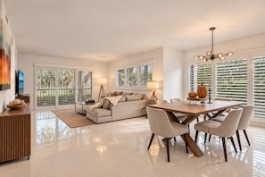 A spacious living area with a sofa, dining table, and chairs, large windows with blinds, and a chandelier.