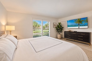 A bedroom with a bed, a nightstand, a lamp, a dresser, a TV, and a window with a view of greenery and a beach.