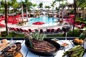 Une table à manger avec un steak, des asperges et un verre de vin rouge, donnant sur un espace piscine agrémenté de parasols rouges et de chaises longues.