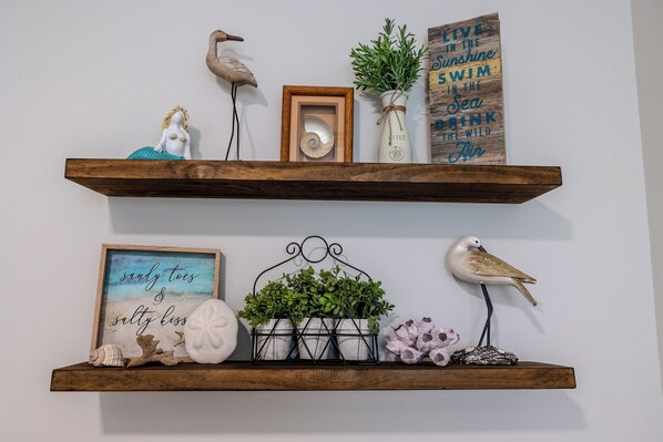 Dos estantes de madera con objetos decorativos, incluyendo una figurita de sirena, un cuadro enmarco, una escultura de pájaro, una planta en maceta y un letrero con una cita motivacional.