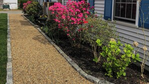 A house with a gravel driveway, a garden with pink flowers, and an American flag. - Immaculate Coastal Retreat! (Tisbury)