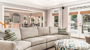 Ein Wohnzimmer mit einem beigefarbenen Ecksofa, einem gemusterten Hocker und einem Essbereich im Hintergrund.
