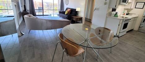 A modern living room with a glass-top table and chairs, a sofa, and a kitchen area with white cabinets and appliances.