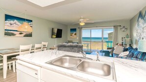 A modern kitchen with a marble countertop and sink, a dining area with chairs, a gray sofa, a ceiling fan, and a view of the beach through large windows. - Ocean Vue - 1BR Surf Condo - Swimming Pool & Ocean Views! (Surf City)