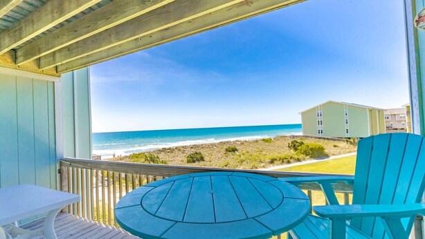 A covered deck with a table and chairs, overlooking a beach and a building. - Ocean Vue - 1BR Surf Condo - Swimming Pool & Ocean Views! (Surf City)