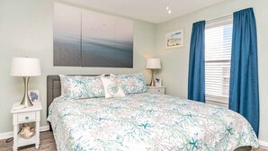 A bedroom with a bed, two bedside tables, a ceiling fan, a window with curtains, and a wall with a painting. - Ocean Vue - 1BR Surf Condo - Swimming Pool & Ocean Views! (Surf City)
