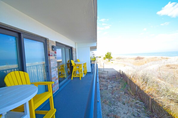 A balcony with yellow chairs and a table, overlooking a beach with a fence and sparse vegetation.