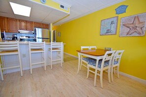A bright yellow kitchen with wooden cabinets, a stainless steel refrigerator, and a dining area with white chairs and a wooden table.