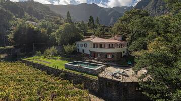 Una casa de dos pisos con piscina, rodeada de abundante vegetación y montañas.
