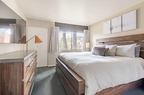 Un dormitorio con una cama grande, un aparador de madera, un escritorio, una ventana con cortinas y vistas a árboles.