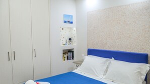 Una cama bien hecha con una colcha azul y almohadas blancas, dos toallas enrolladas sobre la cama, un armario blanco con un estante encima, y una pared con papel tapiz estampado.