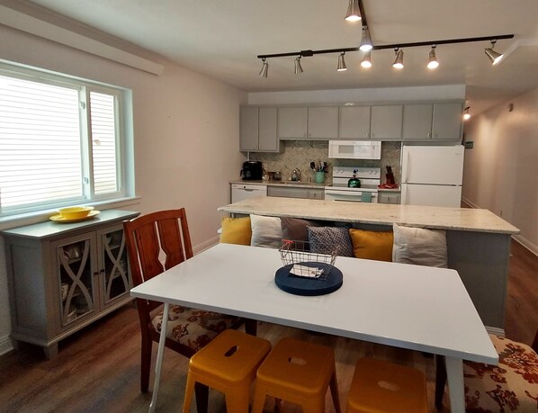 Cozinha compacta com área de refeições, apresentando uma mesa branca, banquetas amarelas e uma janela com persianas.