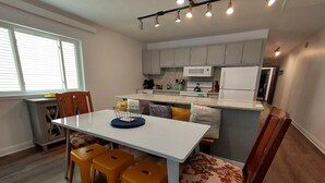 Cozinha compacta com armários brancos, micro-ondas e fogão. Possui um espaço de refeições com mesa branca e quatro banquetas.