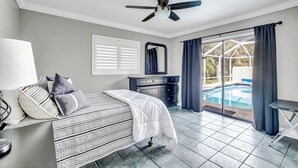 Camera da letto con un letto, una cassettiera, una ventola a soffitto, una finestra con persiane e vista su una piscina.