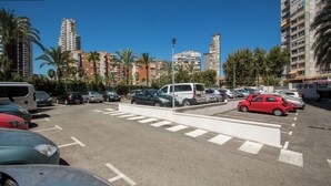 A parking lot with multiple cars and a white van, surrounded by palm trees and tall buildings. - AQUARIUM II 6-3 (Benidorm)