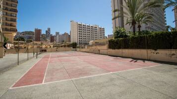 An outdoor tennis court surrounded by high-rise buildings, with a clear sky above.
