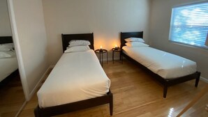 Ein kleines, sauberes Zimmer mit zwei Einzelbetten, einem Nachttisch mit Lampe und einem Fenster mit Jalousien.