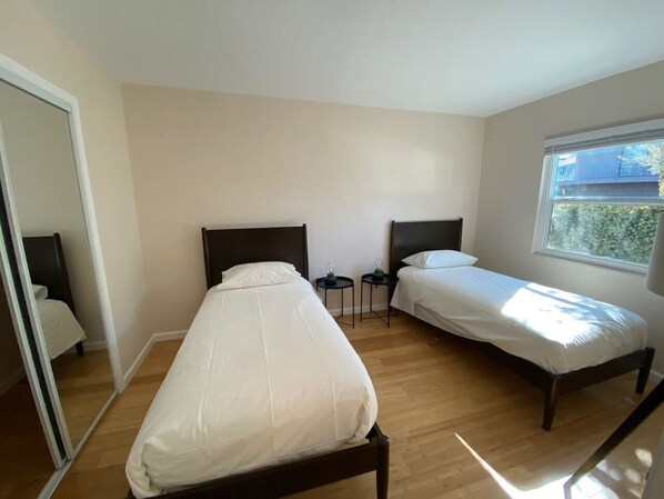 Un dormitorio con dos camas individuales, una mesita de noche con lámpara y una ventana con vistas a edificios.