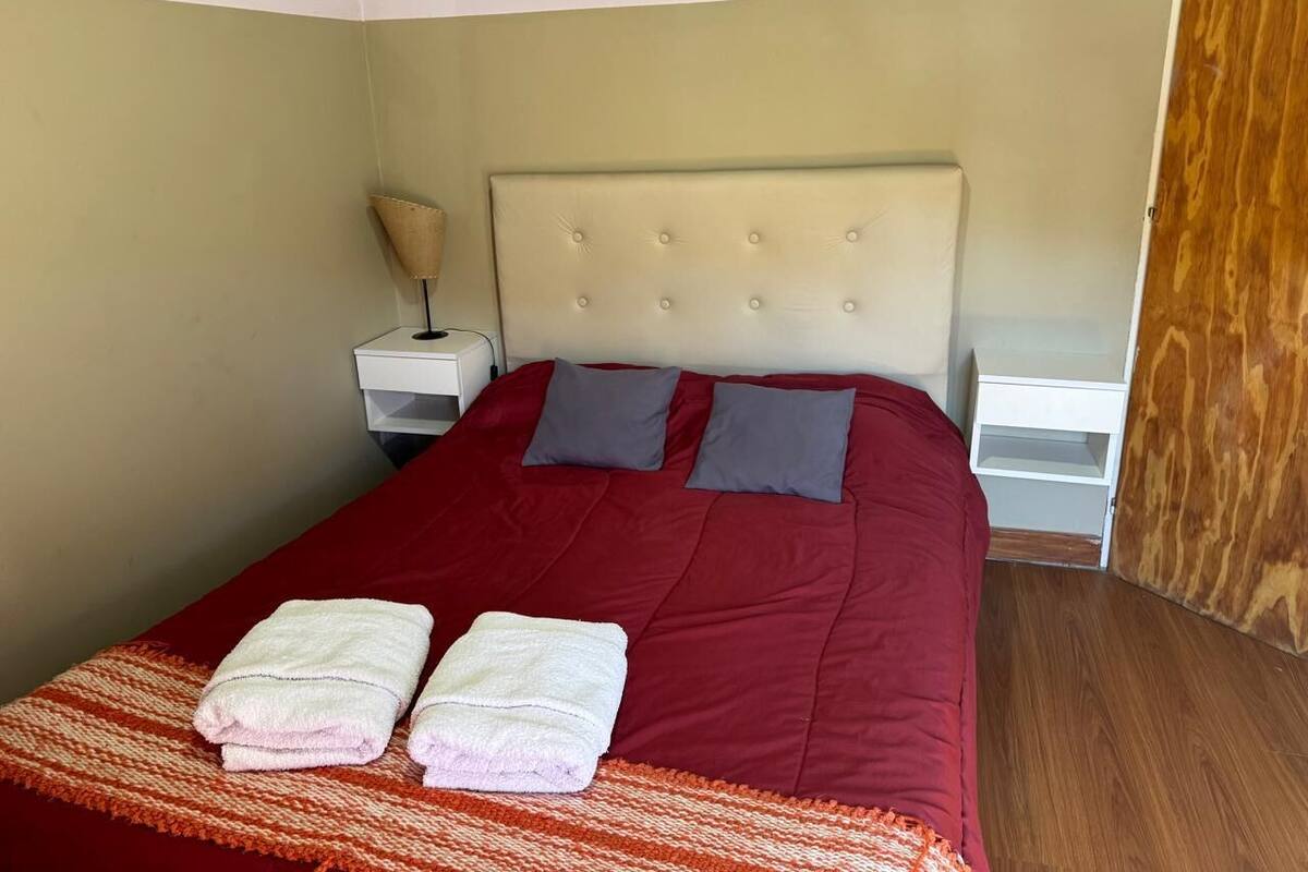 Una cama bien hecha con ropa de cama roja y dos almohadas, una mesita de noche con una lámpara y suelo de madera.