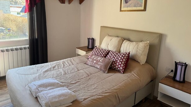 Une chambre avec un grand lit, deux tables de chevet, une fenêtre avec des rideaux, un radiateur et un tableau au mur.