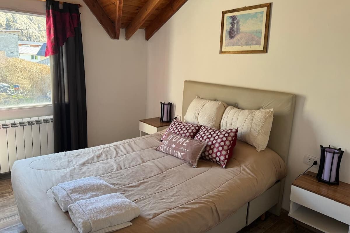 Un dormitorio con una cama grande, dos mesitas de noche, una ventana con cortinas, un radiador y un cuadro en la pared.