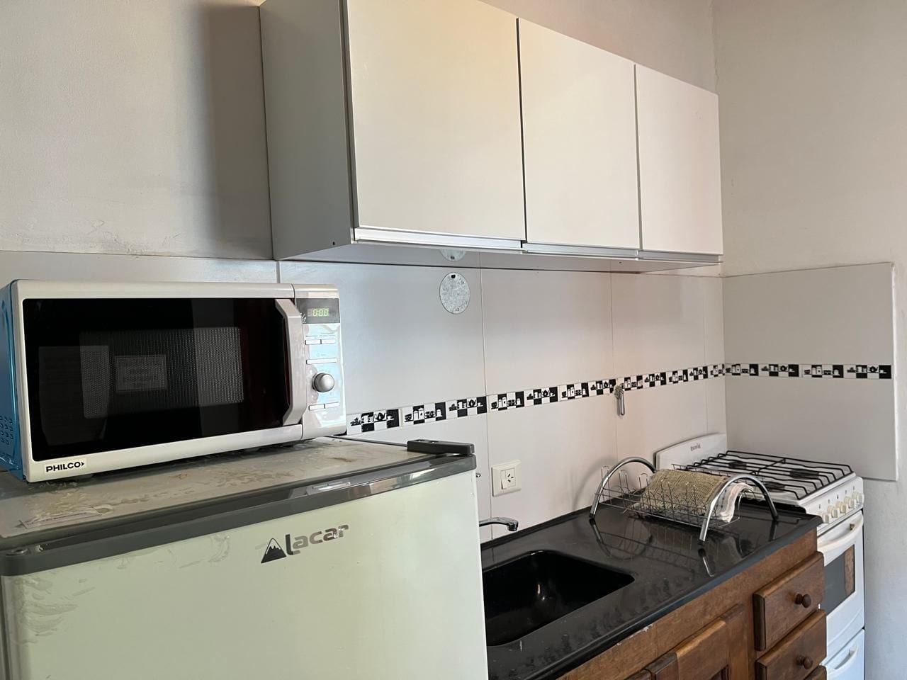 Una cocina compacta con un microondas PHILCO, un refrigerador Lacer, una estufa a gas y una encimera negra.