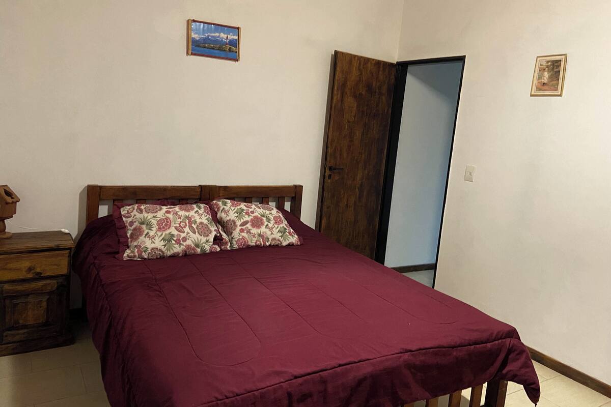 Un dormitorio con estructura de cama de madera, colcha roja y dos almohadas estampadas. Hay una mesita de noche de madera con una lámpara, un aparador de madera y una puerta que da a otra habitación.