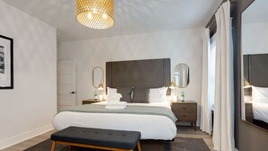 Una habitación de hotel moderna con una cama grande, un banco, una mesita de noche con lámpara y un espejo.