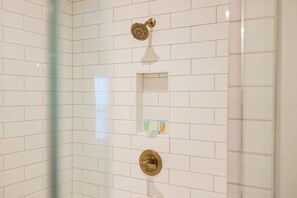 Un baño con azulejos blancos tipo metro, un cabezal de ducha, un espejo y un pequeño estante con artículos de aseo.