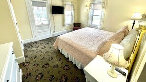 A bedroom with a bed, a chair, a desk with a lamp, and a TV on the wall. - Quaker Inn (Ocean Grove)