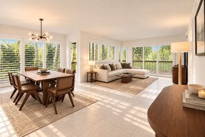 A spacious living area with a dining table, sofa, and a view of greenery through large windows.