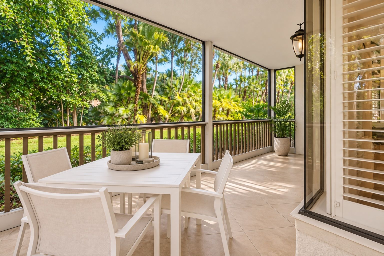 A covered outdoor area with a white table and chairs, surrounded by a glass railing, overlooking a lush garden with palm trees.
